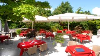 Vue extérieure de la terrasse avec des nappes rouges, entourée d'arbres et de parasols.