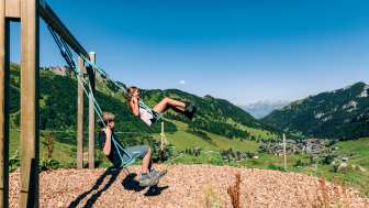 Deux enfants se balancent sur une aire de jeux de montagne avec vue sur le panorama estival de Malbun au Liechtenstein.