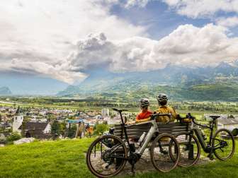 Deux cyclistes sont assis avec leur casque sur un banc au-dessus de la vallée du Rhin et profitent de la vue sur la vallée et les montagnes environnantes - expérience de gravel bike au Liechtenstein