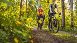 Deux cyclistes portant des casques roulent sur leurs vélos gravel sur un sentier forestier ensoleillé à travers une canopée verte – des vacances actives dans la nature du Liechtenstein
