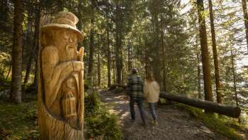 Deux randonneurs observent des figures en bois sculptées sur le « sentier des légendes walsers » un sentier thématique au milieu d'un paysage alpin avec vue sur les montagnes.