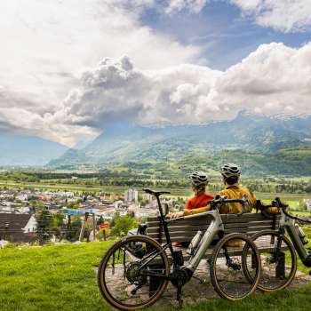 Deux cyclistes sont assis avec leur casque sur un banc au-dessus de la vallée du Rhin et profitent de la vue sur la vallée et les montagnes environnantes - expérience de gravel bike au Liechtenstein