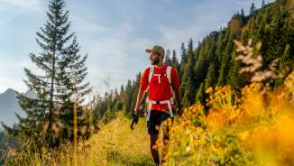 Randonneur en t-shirt rouge sur un chemin de montagne ensoleillé près de Malbun, entouré de fleurs de prairie, de forêts de conifères et d'un décor alpin au Liechtenstein.