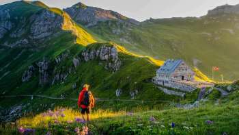 Un randonneur regarde le refuge Pfälzerhütte au Liechtenstein, entouré d'un paysage de montagnes verdoyantes et d'un panorama alpin au crépuscule.