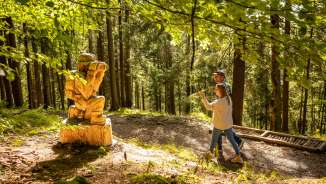 Deux visiteurs découvrent une figure en bois sculptée le long du « sentier des légendes walsers » à Triesenberg, Liechtenstein - sentier de randonnée thématique mystique dans la forêt