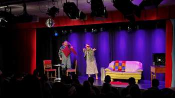 Scène de cabaret au K-BUM au Liechtenstein - scène colorée avec canapé, duo d'acteurs et public dans la salle.