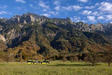 Un bus LIEmobil jaune et noir traverse un paysage de prairies automnales devant de hautes montagnes.