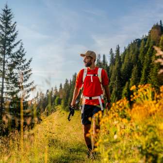 Randonneur en t-shirt rouge sur un chemin de montagne ensoleillé près de Malbun, entouré de fleurs de prairie, de forêts de conifères et d'un décor alpin au Liechtenstein.