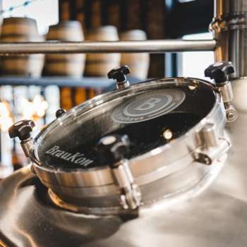 Gros plan d'un hublot rond sur une cuve de brassage brillante avec vue sur la salle de brassage en arrière-plan.