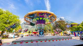 Grand carrousel à chaînes multicolore à la foire de Balzers, plein de visiteurs joyeux devant un décor de montagne sous un ciel bleu.