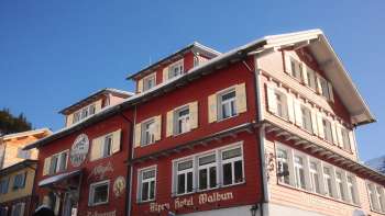 Vue extérieure de l'Alpenhotel Vögeli Malbun avec le bar à élans au rez-de-chaussée - maison de tradition rouge avec une inscription blanche sur fond de ciel bleu.