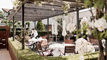 Une terrasse élégante avec des tables blanches, couvertes de serviettes et de verres, sous un pavillon en bois entouré d'une verdure luxuriante.