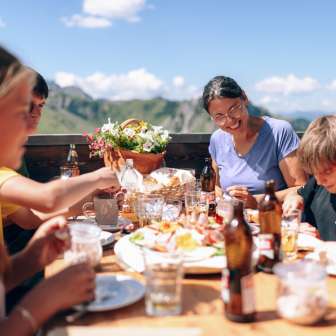 Famille déjeunant ensemble sur une terrasse ensoleillée à Sarreis, entourée de montagnes et de spécialités régionales.