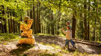 Deux randonneurs passent devant une statue en bois sur le « sentier des légendes walsers »