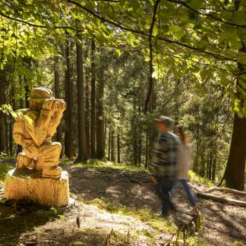 Des randonneurs passent devant un personnage sculpté avec des jumelles au « sentier des légendes walsers »