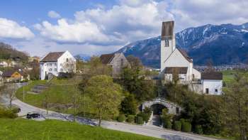 Vue panoramique sur le « Kirchhügel » (colline de l'église) de Bendern avec son église historique, entourée de chemins bien entretenus, d'arbres et d'une architecture traditionnelle dans un décor de montagne alpin.