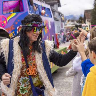 Hippie déguisée lors du carnaval des enfants à Mauren, high-five avec des enfants devant un char coloré.