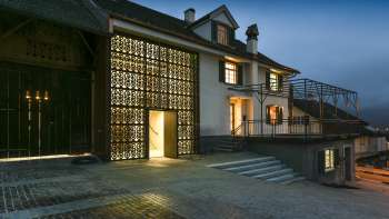Maison de la culture Rössle à Mauren éclairée de façon moderne au crépuscule, avec façade en verre, annexe historique du bâtiment et esplanade pavée.
