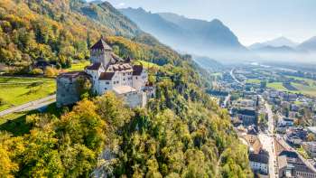 Vue aérienne du château de Vaduz sur une colline au-dessus de la ville, avec panorama sur les Alpes et vue sur la vallée du Rhin par temps clair.
