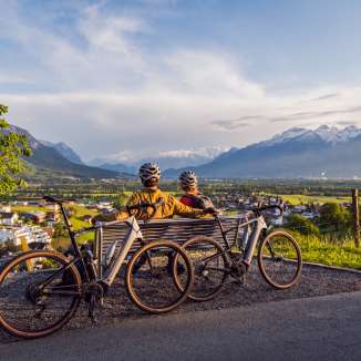Deux cyclistes sont assis avec leur casque sur un banc au-dessus de la vallée du Rhin et profitent du coucher de soleil avec vue sur les Alpes - point fort du cyclotourisme au Liechtenstein