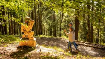 Deux randonneurs passent devant une statue en bois sur le « sentier des légendes walsers »