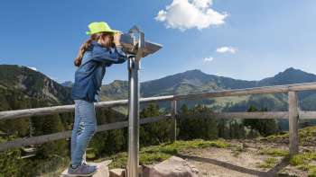 Fillette au chapeau vert regardant à travers un télescope sur le sentier des chercheurs de Malbun avec vue panoramique sur les Alpes liechtensteinoises.