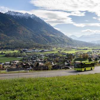 Le bus vert LIEmobil emprunte une route sinueuse au-dessus du Liechtenstein avec vue sur la vallée du Rhin et les Alpes environnantes.