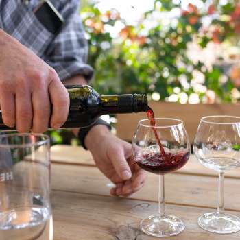 Un verre de vin rouge est versé d'une bouteille dans un verre à vin, avec des plantes en fleurs en arrière-plan.