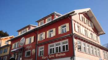 Vue extérieure de l'Alpenhotel Vögeli Malbun avec le bar à élans au rez-de-chaussée - maison de tradition rouge avec une inscription blanche sur fond de ciel bleu.
