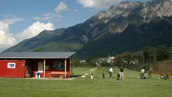 Des golfeurs sur le practice au Liechtenstein avec vue sur des prairies verdoyantes, un club-house rouge et le paysage montagneux environnant sous un ciel bleu.