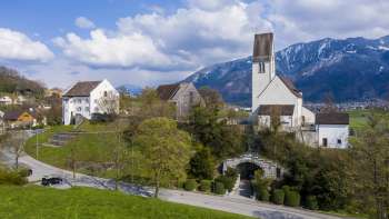 Vue panoramique sur le « Kirchhügel » (colline de l'église) de Bendern avec son église historique, entourée de chemins bien entretenus, d'arbres et d'une architecture traditionnelle dans un décor de montagne alpin.