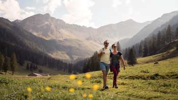 Deux personnes se promènent dans une vaste prairie alpine avec vue sur les montagnes environnantes de l'alpage de Valüna par temps ensoleillé.
