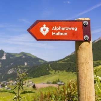Panneau rouge avec l'inscription "parcours du cœur alpin Malbun" devant un pittoresque décor de montagnes et une prairie d'été au Liechtenstein.