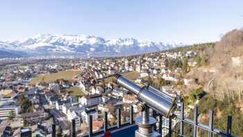 Télescope au point de vue du Schlossweg Känzile avec vue sur Vaduz et le panorama alpin enneigé en arrière-plan.