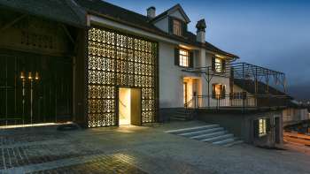 Maison de la culture Rössle à Mauren éclairée de façon moderne au crépuscule, avec façade en verre, annexe historique du bâtiment et esplanade pavée.