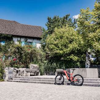 vélo électrique rouge devant un bâtiment historique du Liechtenstein, entouré d'arbres et de fontaines, point de départ idéal pour une balade à vélo à travers le pays.
