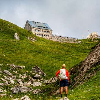 Randonneur s'approchant du refuge Pfälzer sur un versant verdoyant du Liechtenstein, encadré de rochers et de prairies