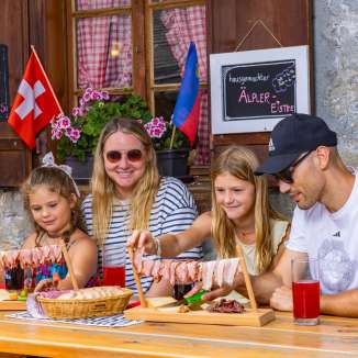 Famille assise sur un alpage ensoleillé du Liechtenstein, savourant une collation traditionnelle avec des spécialités régionales et un panorama de montagne.