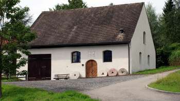 Moulin traditionnel à Eschen avec une façade blanche, des volets sombres et un ancien ensemble de meules devant l'entrée - entouré de prairies verdoyantes et d'arbres.