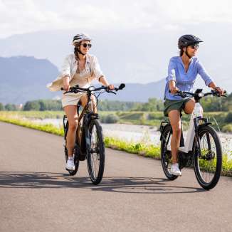 Deux femmes sur des vélos électriques sur la piste cyclable du Rhin près du Liechtenstein, avec le fleuve, un paysage verdoyant et des montagnes en arrière-plan.