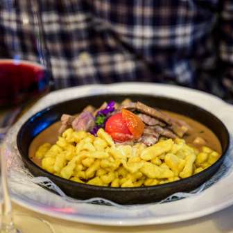 Spätzle savoureux avec ragoût de bœuf, garnis de tomate, servis dans une poêle - accompagnés d'un verre de vin rouge sur une table dressée.