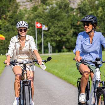 Deux femmes sur des vélos électriques lors d'un tour estival avec les drapeaux du Liechtenstein, de la Suisse et de l'Autriche en arrière-plan