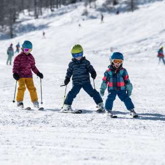 Trois enfants apprennent à skier dans le domaine skiable familial de Malbun au Liechtenstein, sous le soleil et la poudreuse.