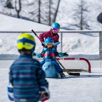 Des enfants jouent avec un casque et une crosse de hockey sur la patinoire de Malbun, un enfant est assis sur un taureau auxiliaire de patinage devant une porte.