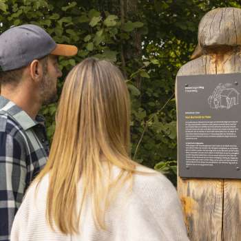 Deux personnes lisent un panneau d'information avec une légende au « sentier des légendes walsers » Triesenberg, sur une figure en bois sculptée.
