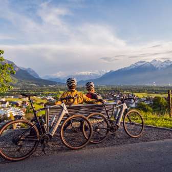 Deux cyclistes sont assis avec leur casque sur un banc au-dessus de la vallée du Rhin et profitent du coucher de soleil avec vue sur les Alpes - point fort du cyclotourisme au Liechtenstein