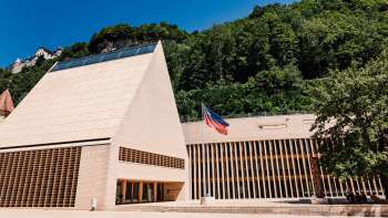 Bâtiment moderne du Landtag à Vaduz avec un pignon pointu, devant le drapeau du Liechtenstein et entouré d'un décor de collines verdoyantes.