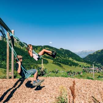 Deux enfants se balancent sur une aire de jeux de montagne avec vue sur le panorama estival de Malbun au Liechtenstein.