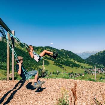 Deux enfants se balancent sur une aire de jeux de montagne avec vue sur le panorama estival de Malbun au Liechtenstein.