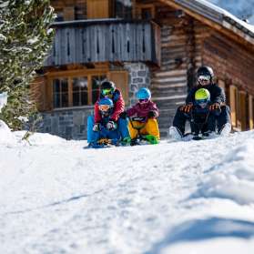 Deux adultes et trois enfants assis sur des luges entament ensemble une descente devant un chalet de montagne enneigé à Malbun.